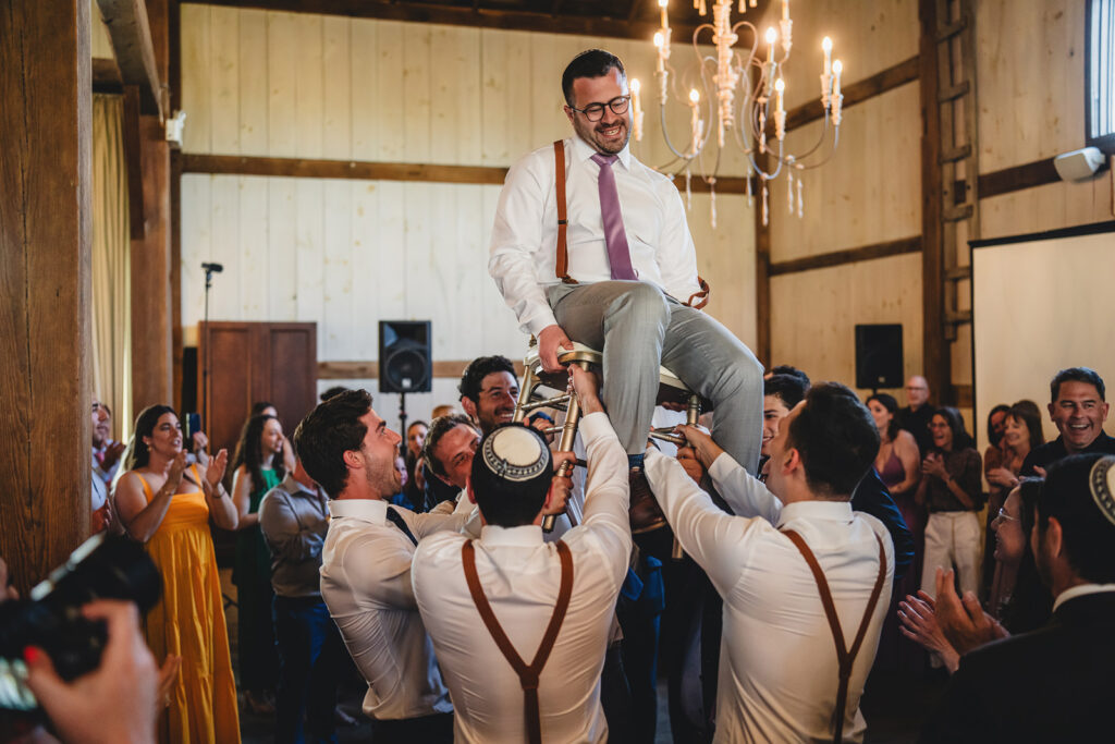 A groom lifted high on a chair at The Earth to Table Farm wedding venue